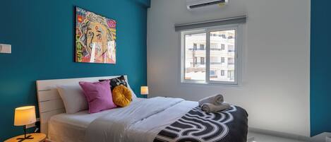 A bedroom with a bed, a nightstand, a lamp, a window, and a wall-mounted air conditioner.