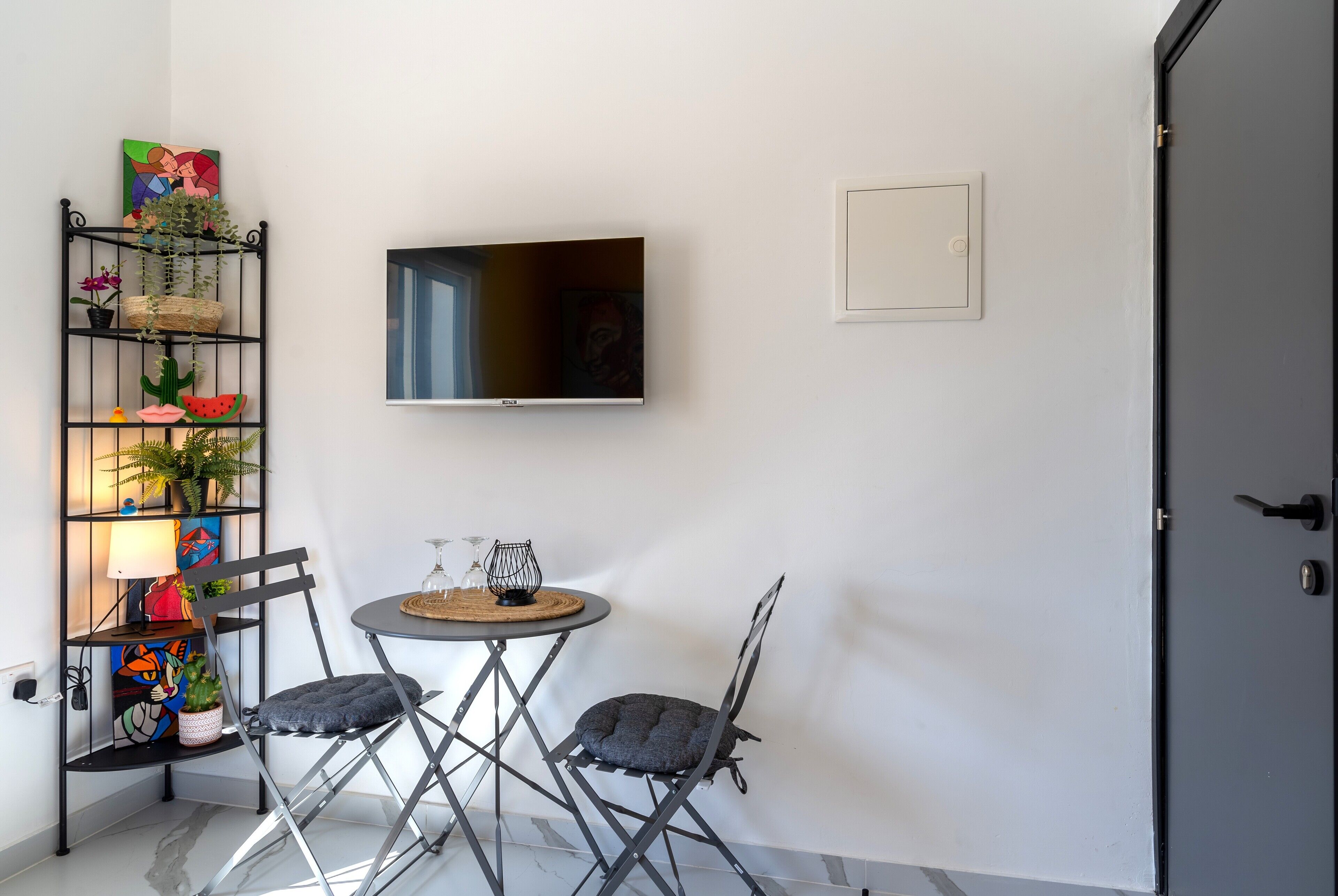 Een compacte leefruimte met een flatscreen televisie aan de muur, een klein rond tafeltje met een kaars, twee opklapbare stoelen en een decoratieve plank met planten en kunstwerk.