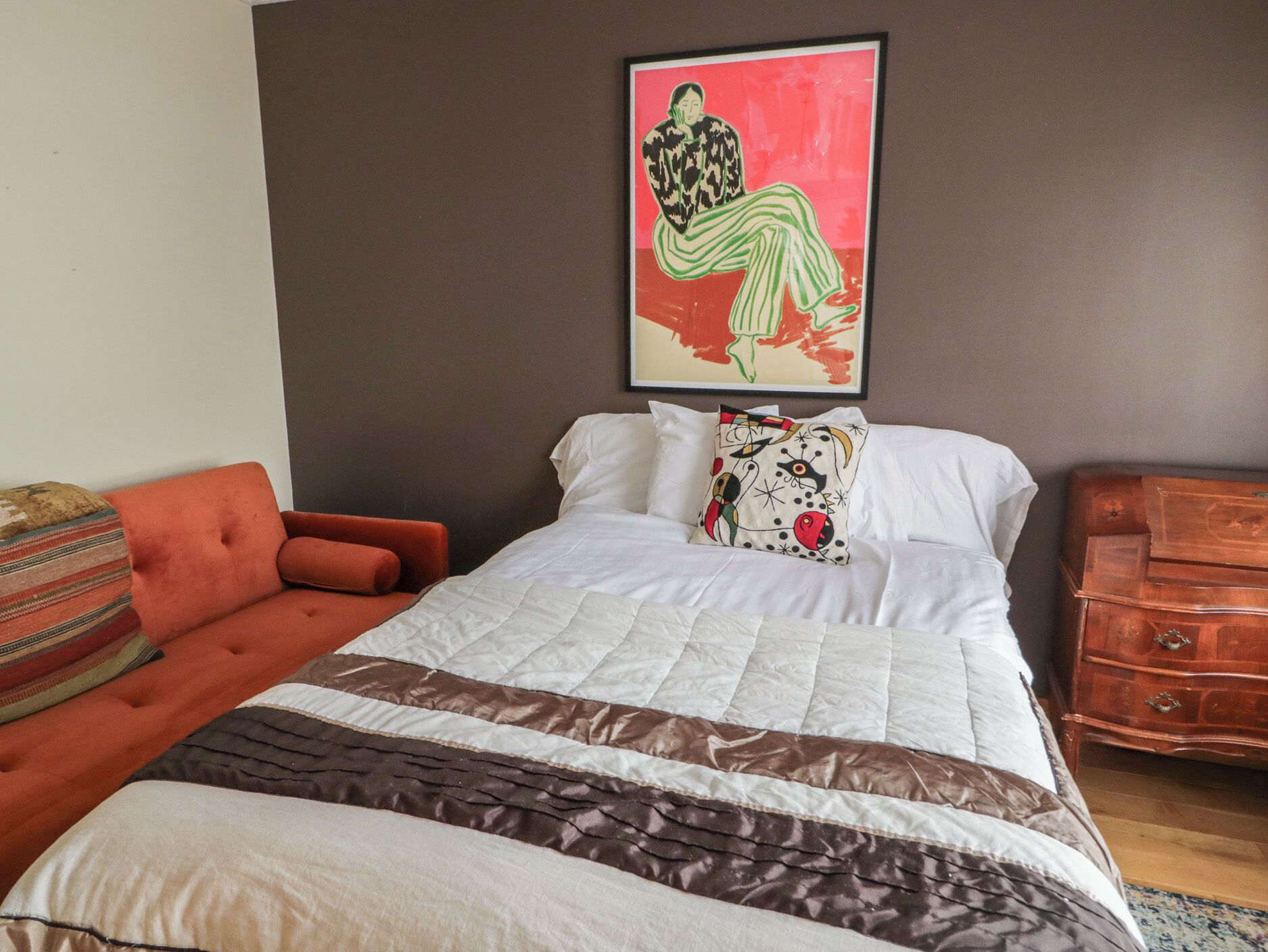 A bedroom with a bed, a red armchair, a wooden dresser, and a painting on the wall.