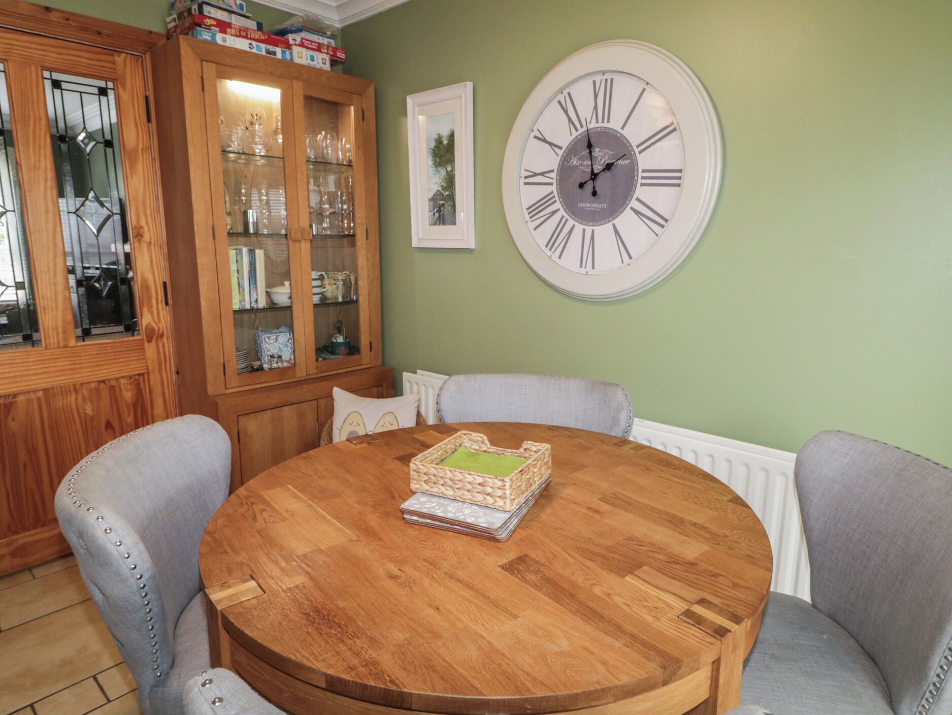 Une table ronde en bois, sur laquelle sont posés un panier tressé et un tableau encadré, entourée de chaises grises recouvertes de tissu.