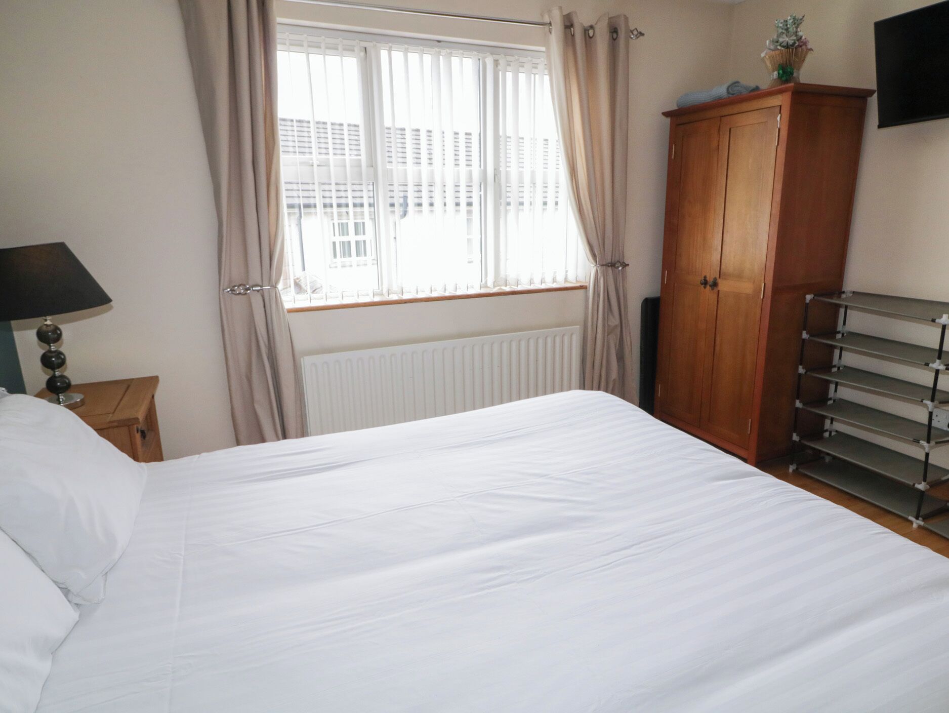 A bedroom with a bed, a radiator, a wooden wardrobe, a lamp, and a window with curtains.