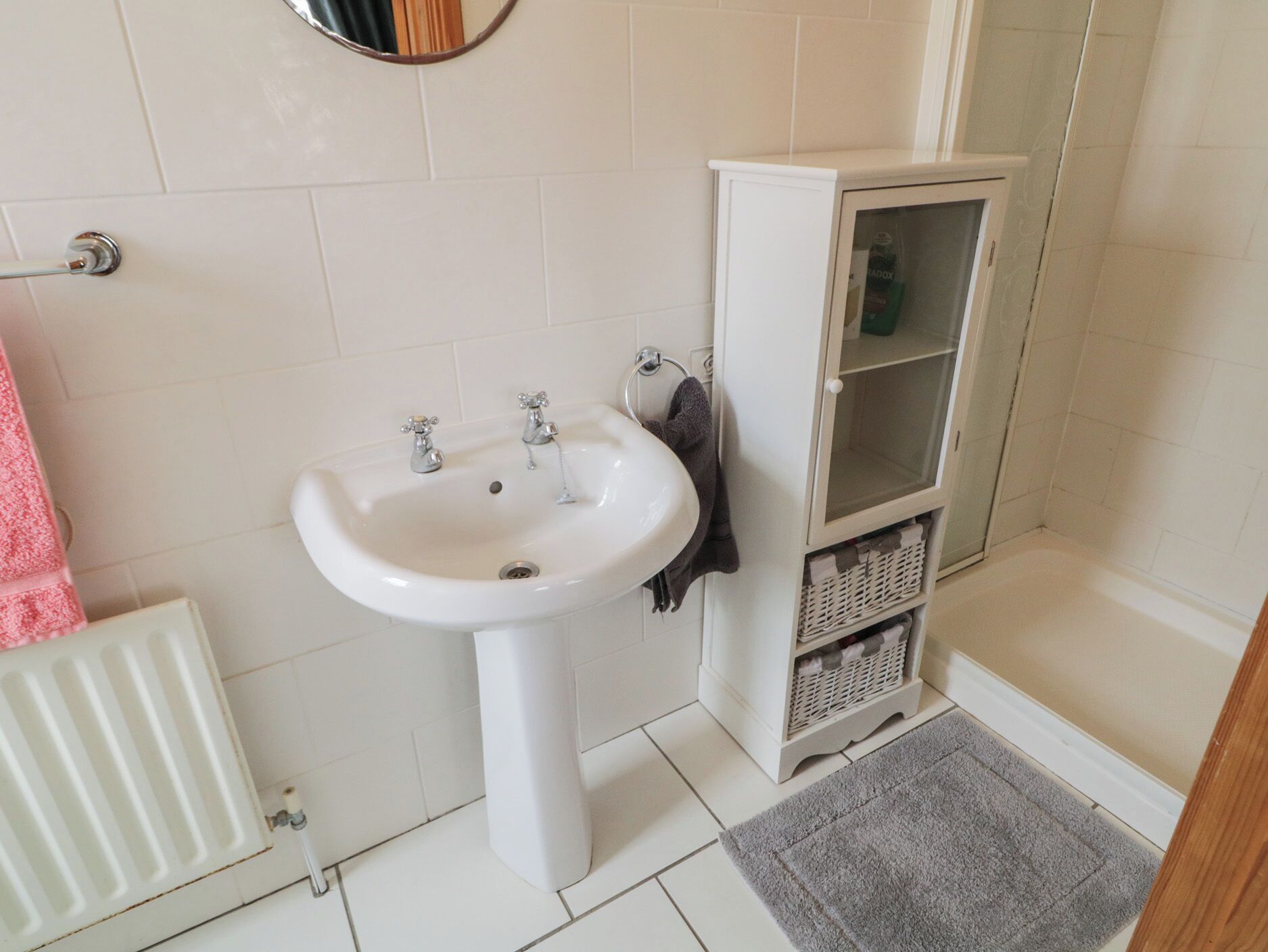 A bathroom with a white sink, a mirror, a towel rack, a radiator, and a cabinet with a glass door.