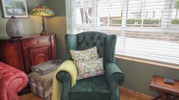 A cozy room with a green armchair, a wooden dresser, a lamp, and a window with blinds.