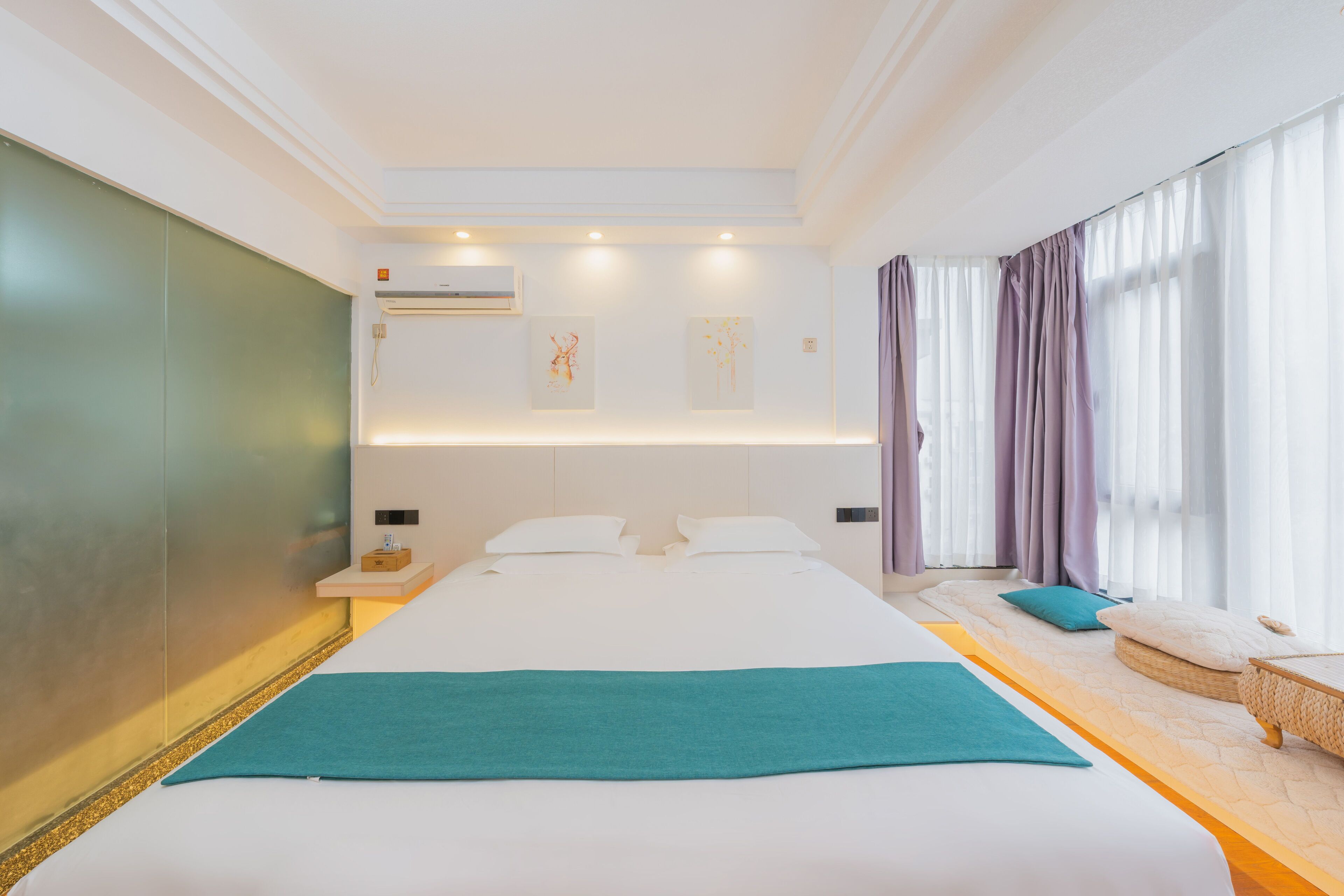A modern hotel room with a large bed, a bedside table, a wall-mounted air conditioner, and a window with curtains.