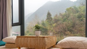Una habitación con un ventanal grande, un reposapiés de mimbre, una planta en maceta y vistas a un paisaje montañoso.