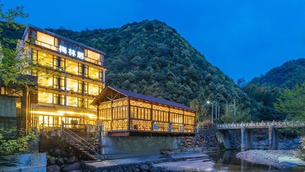 Un edificio de dos pisos con fachada de madera y ventanas amplias, ubicado junto a un río con un puente al fondo.