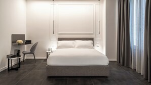 Ein modernes Hotelzimmer mit einem großen Bett, einem Schreibtisch mit Stuhl, einem kleinen Tisch mit Blumenvase und einem Fenster mit Vorhängen.