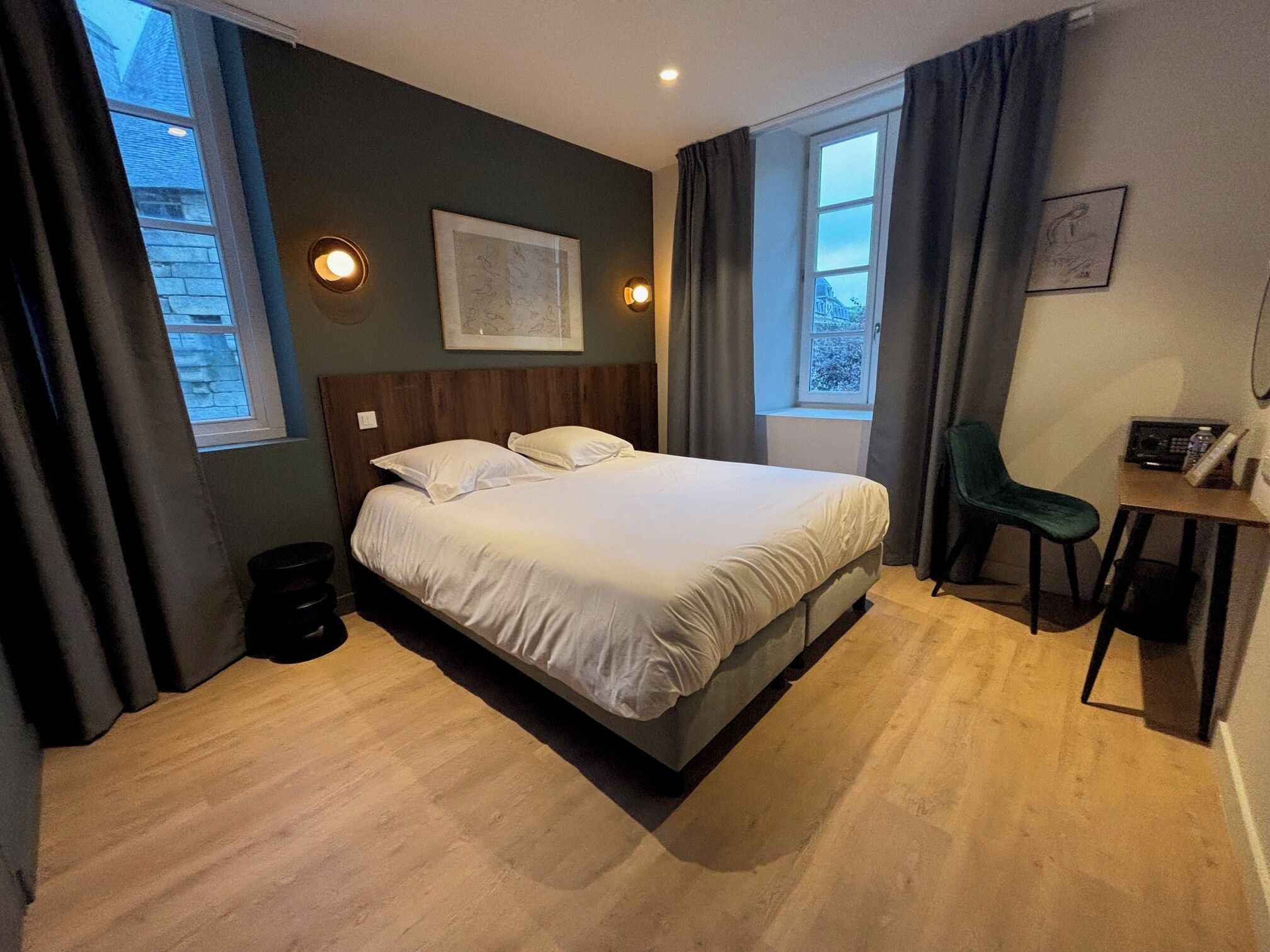 Quarto de hotel com uma cama grande, uma escrivaninha e duas janelas.