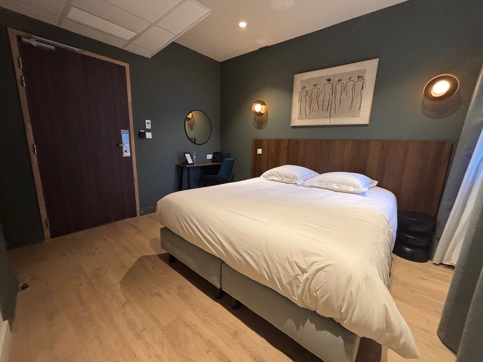 A bedroom with a large bed, a desk, and a chair. There is a door on the left and a wall-mounted light fixture on the right.