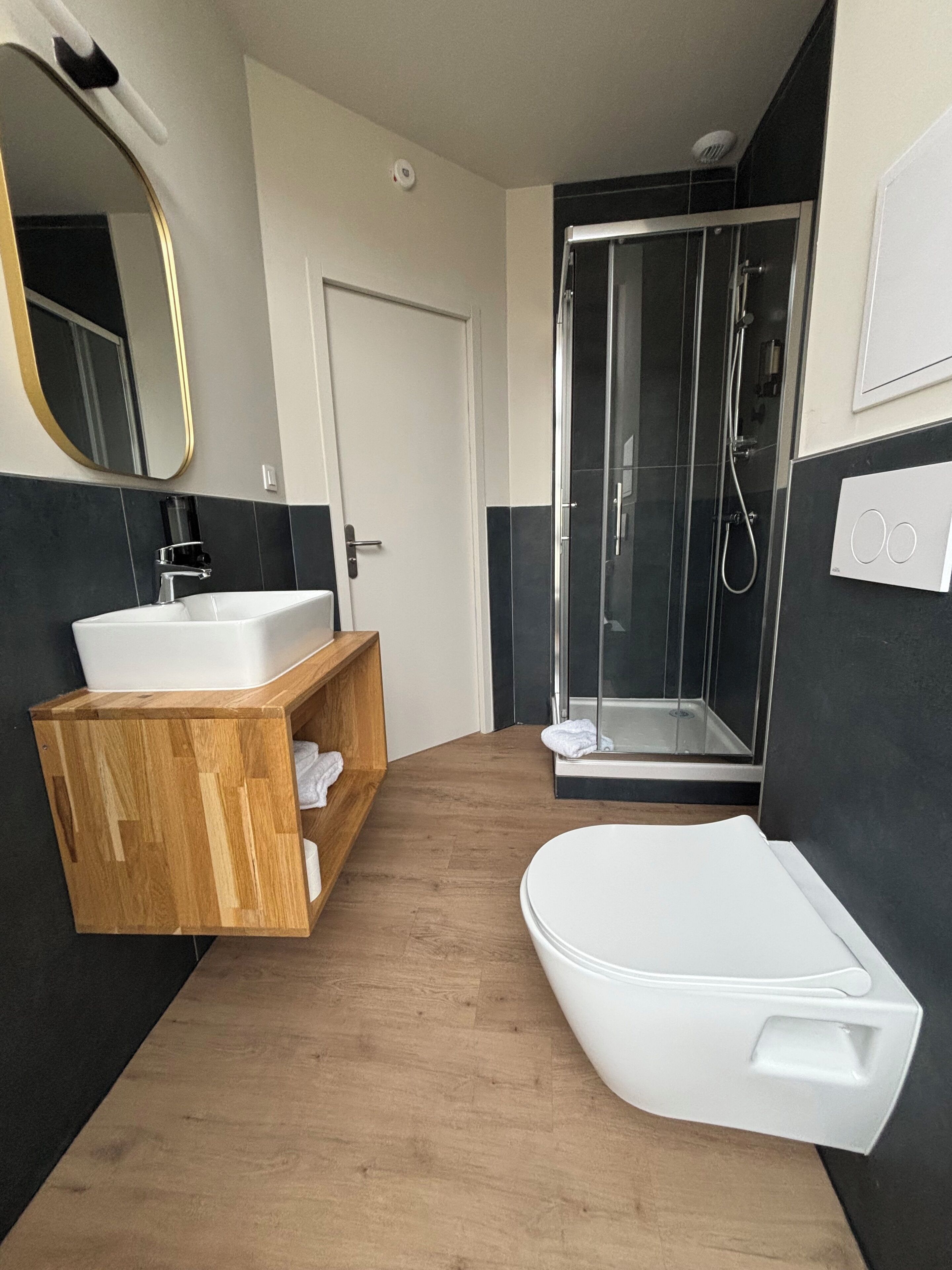 Un baño moderno con tocador de madera, un lavamanos blanco, inodoro y ducha con mampara de vidrio.