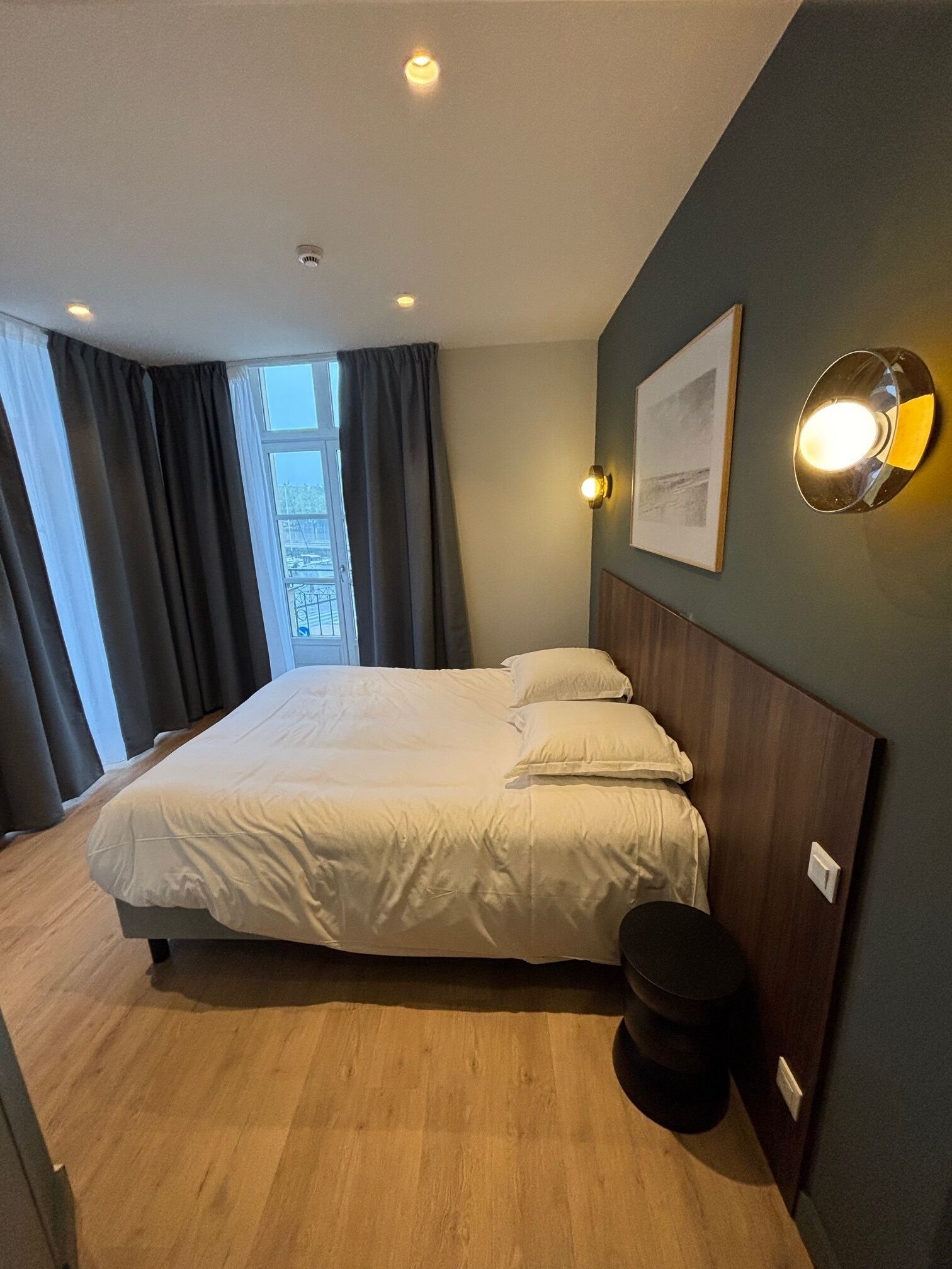 A modern bedroom with a large bed, a bedside table, a wall-mounted light, and a window with curtains.