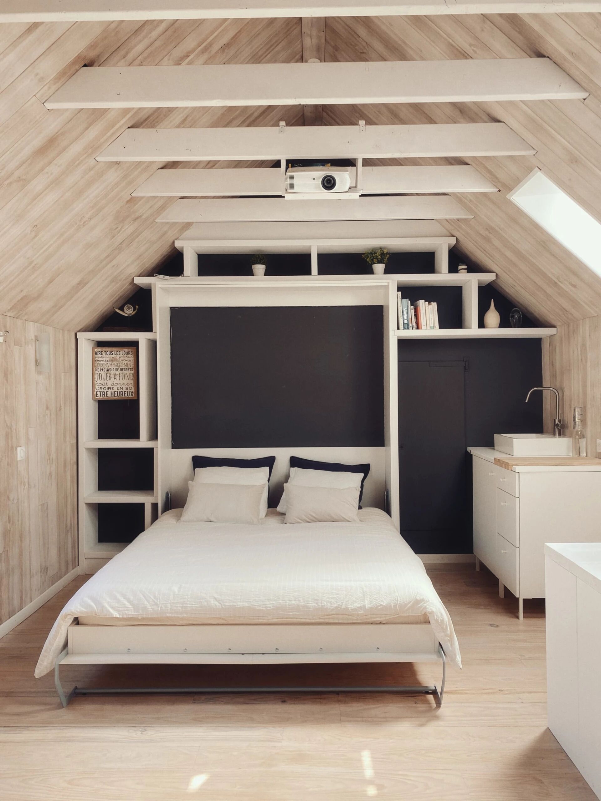 A bedroom with a sloped ceiling, a bed, a nightstand, a dresser, and built-in shelving with books and decorative items.