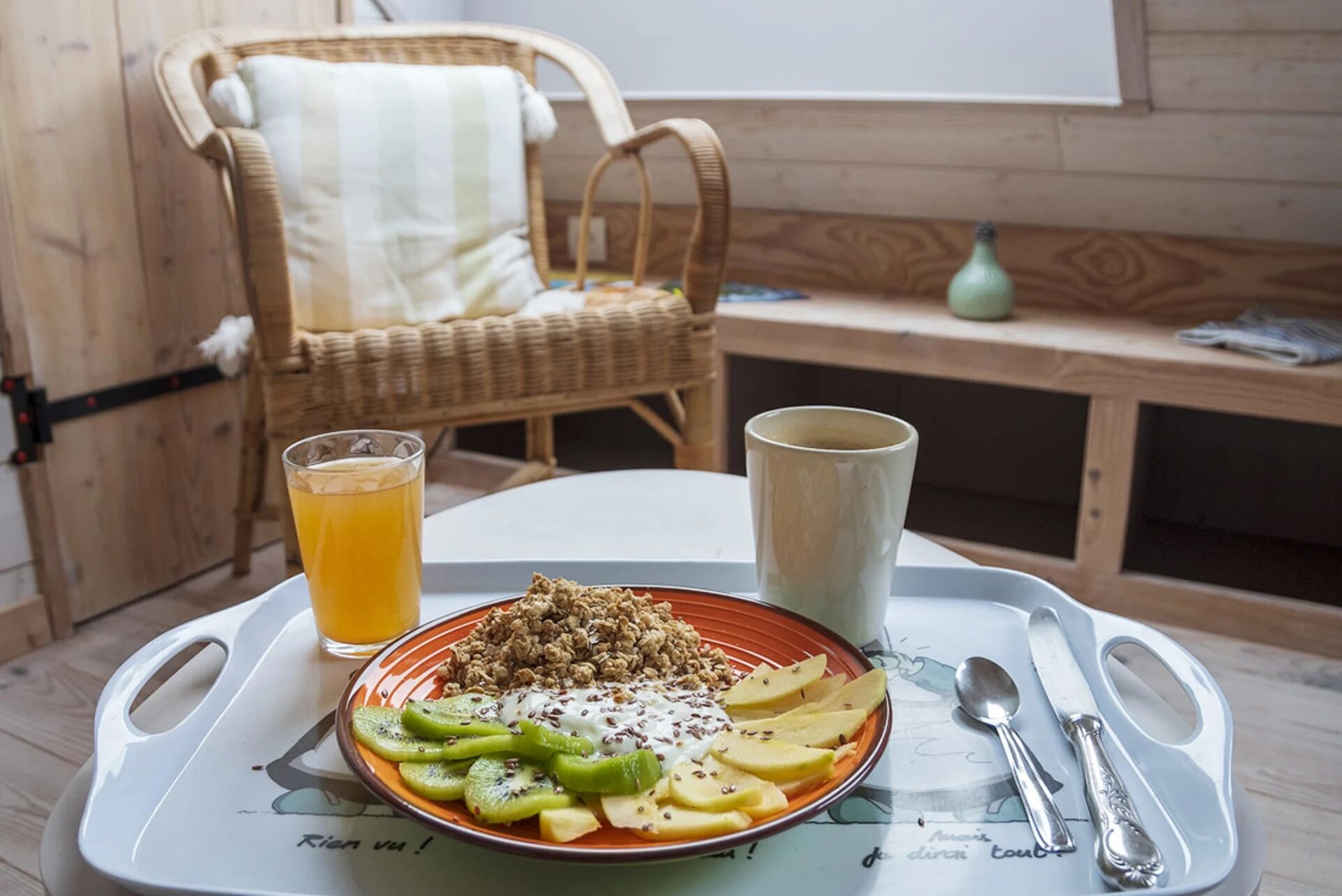 Una bandeja de desayuno con cereal, yogur y fruta sobre una mesa, un vaso de zumo de naranja y una silla al fondo.