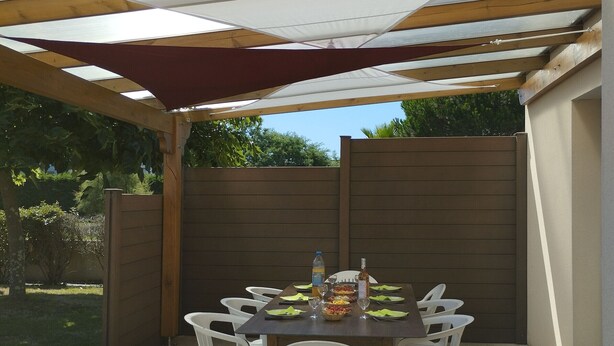 An outdoor patio with a table set for dining, surrounded by white plastic chairs, under a shaded area with a wooden ceiling.