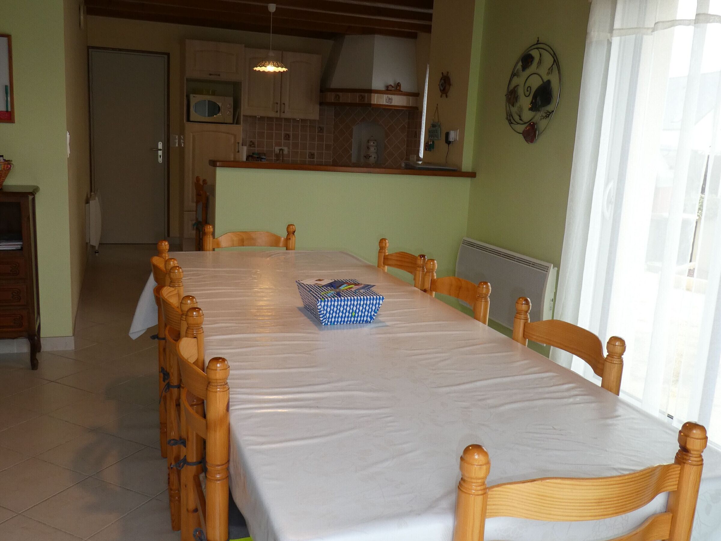 Un comedor con una mesa larga, sillas de madera, un microondas y una cocina al fondo.
