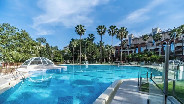 Une piscine entourée d’une structure en verre, bordée de palmiers et d’un bâtiment doté de balcons.