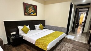 A modern hotel room with a large bed, a nightstand, a painting on the wall, and a view into the hallway. - VR RESIDENCY (Gurugram)