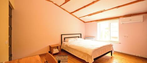 A bedroom with a wooden floor, a bed with white bedding, a wooden bench, a small table, and a window with daylight.