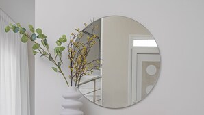 A minimalist room with a white wall, a round mirror, a white vase with branches, and a wooden shelf.