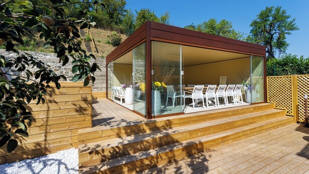 Un espacio de estar al aire libre cubierto con una terraza de madera, paredes de vidrio y sillas blancas.