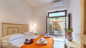 Habitación de hotel con una cama, una mesita con tetera y tazas, un florero con flores, y vistas a un muro de piedra y una zona de estar al aire libre.