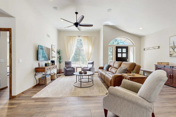 Un salon spacieux doté d’un ventilateur de plafond, d’une grande fenêtre avec des rideaux, d’un canapé en cuir marron, d’un fauteuil gris et d’une table basse.