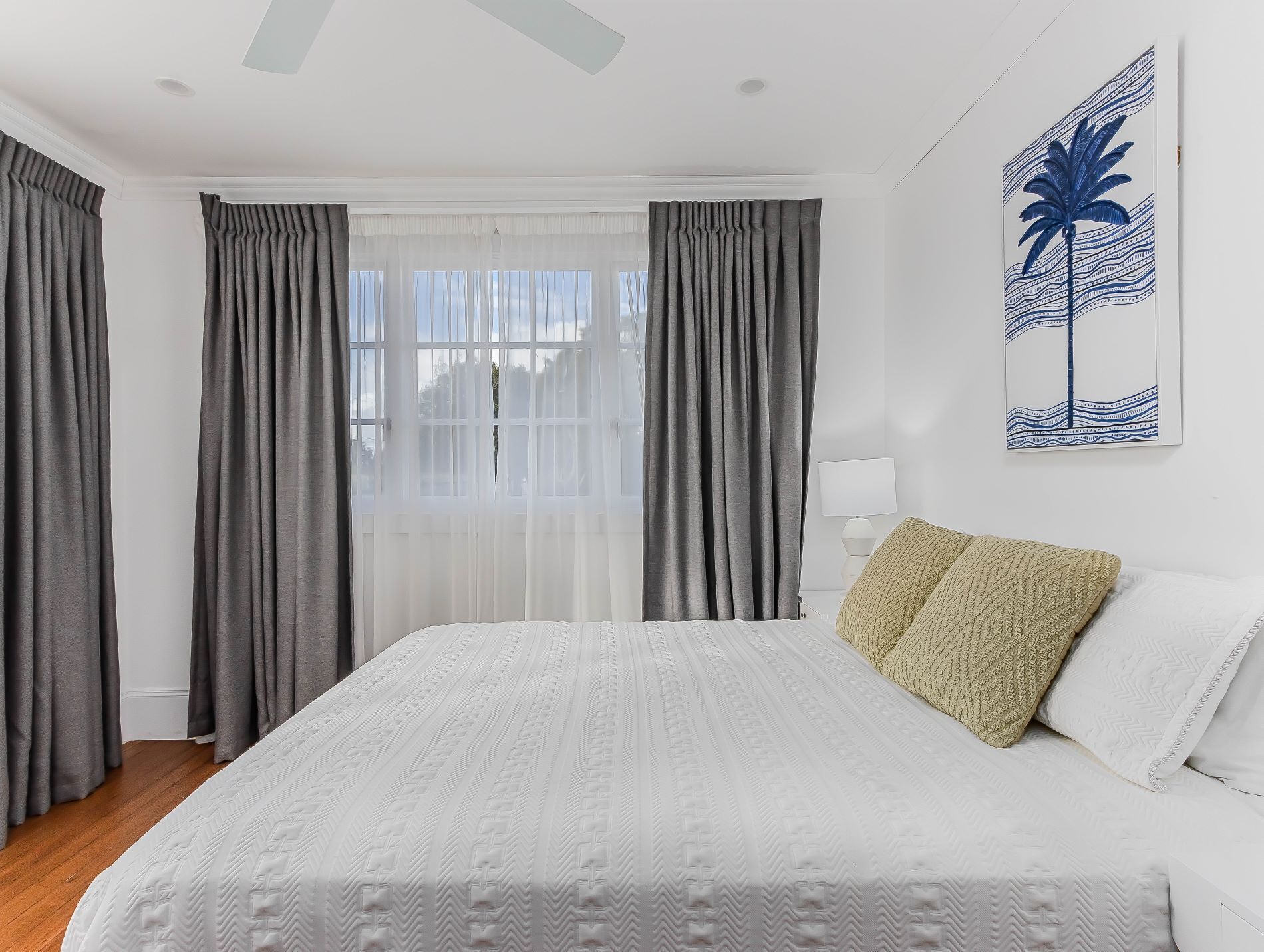 A bedroom with a large bed, grey curtains, a white wall, and a painting of a palm tree.
