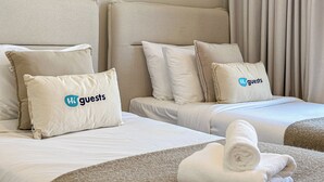 A hotel room with two beds, each with a headboard, white bedding, and pillows with the 'HK guests' logo. There are rolled towels on the bed. - HiGuests - La Rive Classy 2BR & Balcony (Dubai)