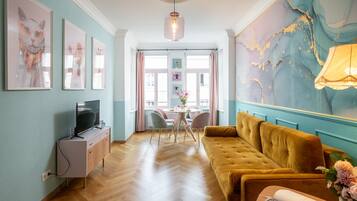 Ein Zimmer mit Holzboden, einem gelben Sofa, einem Fernseher auf einem Schrank und einem großen abstrakten Gemälde an der Wand.