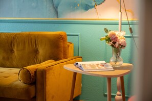 A room with a yellow sofa, a small round table with a magazine, and a vase with flowers.
