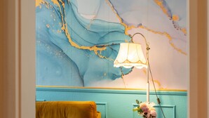 Una habitación con un sofá amarillo, una mesita redonda con una lámpara y flores, y una pared azul verdoso con un diseño que simula mármol.