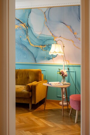 Ein Zimmer mit einem gelben Sofa, einem kleinen runden Tisch mit Lampe und Blumen, sowie einer türkisblauen Wand mit marmorartigem Muster.
