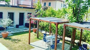 Um pátio com pérgola de madeira, mobiliário de exterior, plantas em vasos e um edifício com varandas ao fundo.