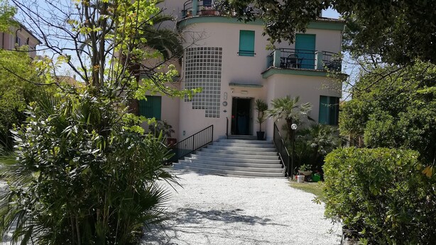 Um edifício com um caminho de cascalho que leva a uma porta, rodeado de vegetação e céu claro.