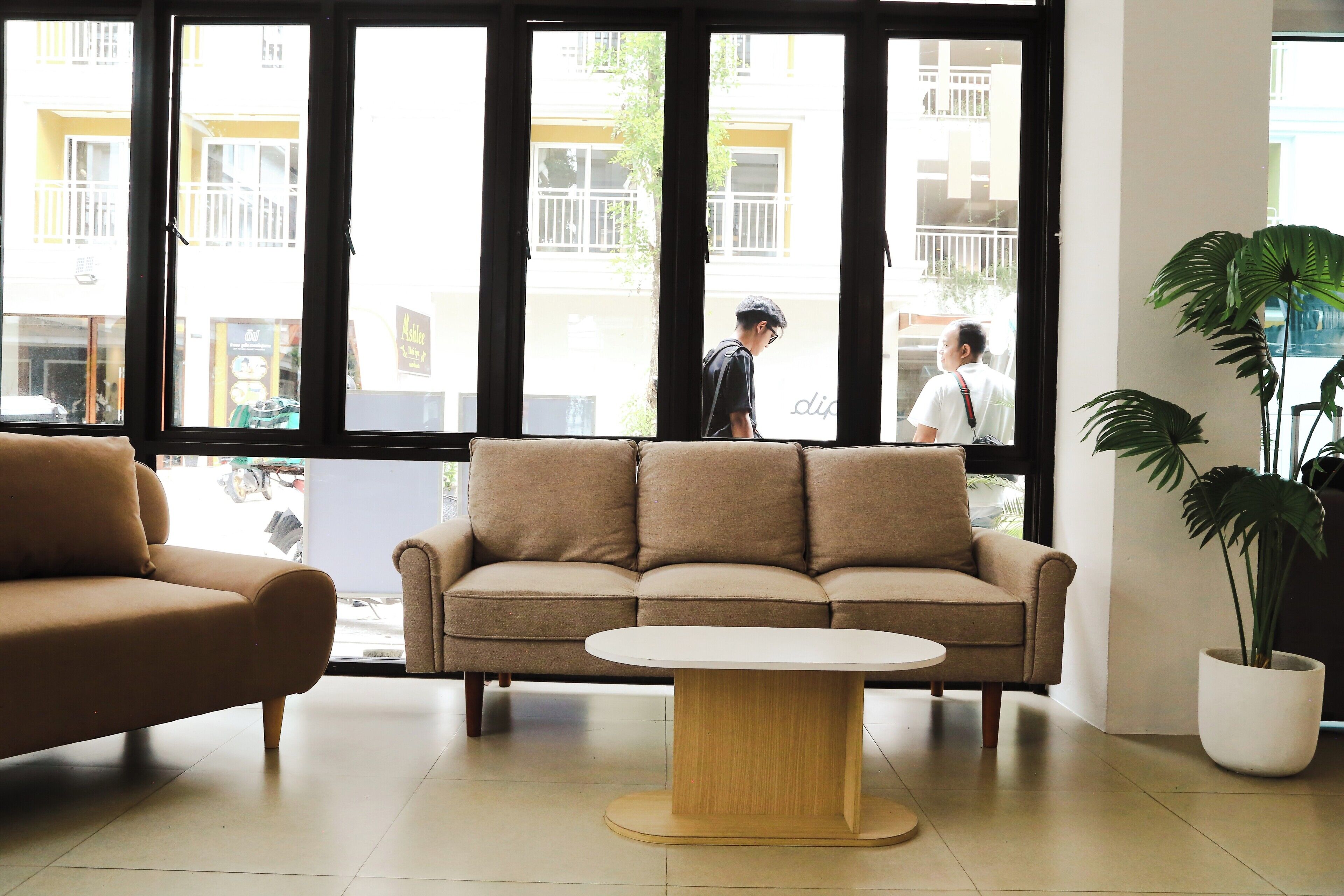 A modern interior with a beige sofa, a round wooden coffee table, and large windows offering a view of the street outside.