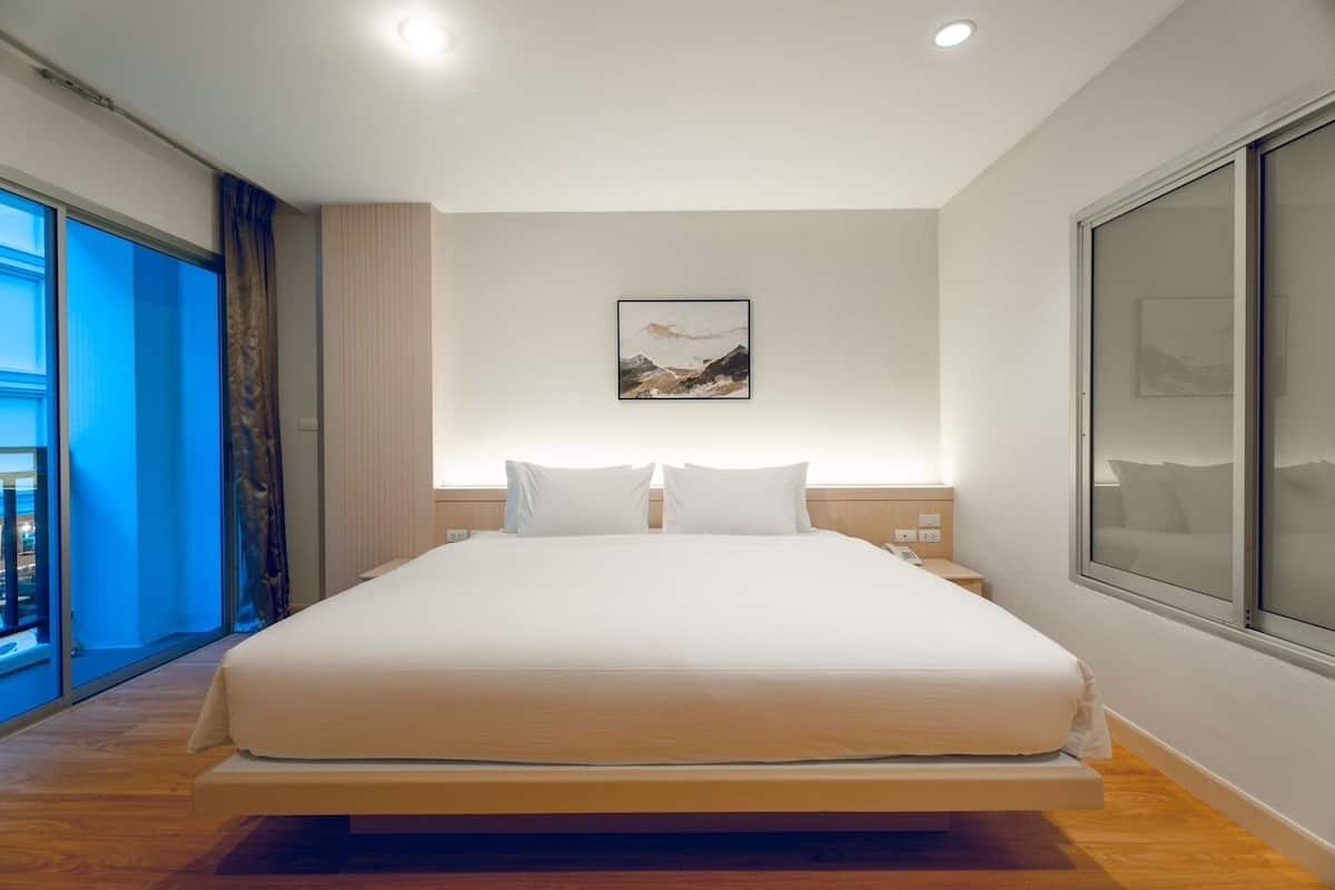 Ein modernes Hotelzimmer mit einem großen Bett, einem Nachttisch mit Lampe, einem großen Fenster mit Aussicht und einem Bild an der Wand.