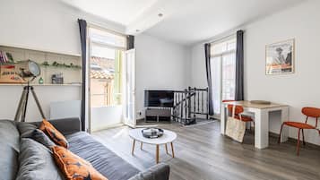Un salon moderne avec un canapé, une table basse et un coin repas avec des chaises. On y trouve également un téléviseur, un escalier et une fenêtre avec des rideaux.