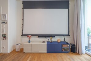 Un salon moderne avec un grand téléviseur à écran plat fixé au mur, un meuble bleu orné d’objets décoratifs, une petite poubelle et un aspirateur posés sur le sol.