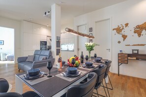 A modern dining area with a long table set for six, surrounded by black chairs. A world map decor is visible on the wall, and a kitchen area is in the background.