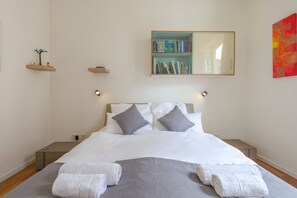 Un lit bien fait, avec des draps blancs, deux oreillers et des serviettes enroulées, posés sur un couvre-lit gris.