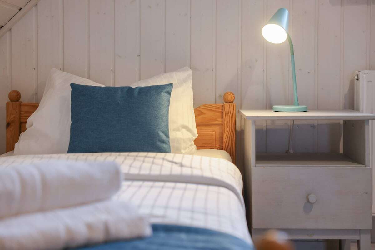Cama bem arrumada com um travesseiro azul, um travesseiro branco e um cobertor branco. Tem uma mesinha de cabeceira branca com uma luminária em cima.