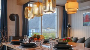 Une table à manger dressée avec des assiettes, des verres et un saladier rempli de fruits, le tout éclairé par des lampes suspendues.