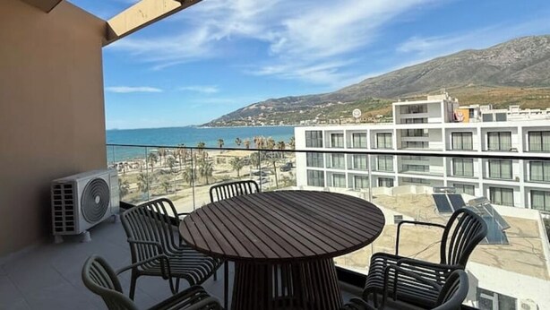 A balcony with a round table and chairs, overlooking a beach and a mountainous landscape.