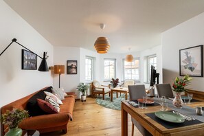 Una sala moderna con piso de madera, un sofá, una mesa de comedor para dos personas y un ventanal que ofrece luz natural.