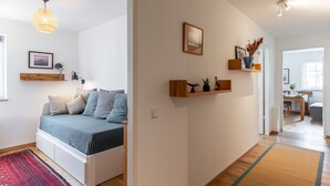 A modern bedroom with a bed, bedside table, and a red rug. There is a hallway leading to a kitchen area. - 3 rooms apartment- 4 people (Leipzig)