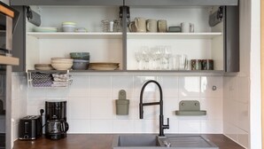 Una cocina con un lavamanos de acero inoxidable, una cafetera y diferentes platos y vasos en estantes.