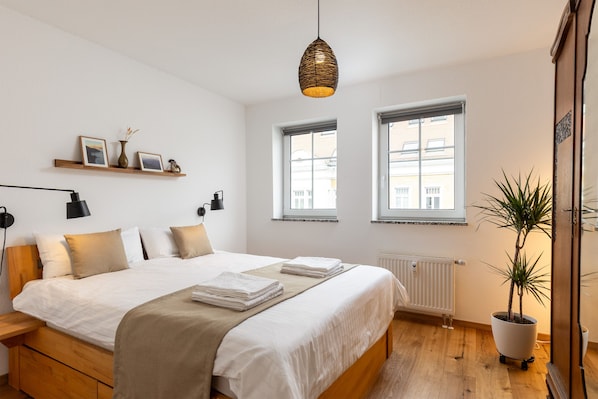 A modern bedroom with a large bed, bedside tables, a hanging light fixture, a potted plant, and windows.