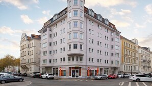 A multi-story building with a corner entrance, multiple windows, and a sign indicating a business on the ground floor. - 3 rooms apartment- 4 people (Leipzig)