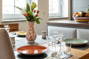 A dining table set with plates, glasses, and cutlery, a vase with flowers, and a fruit bowl on the counter.