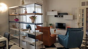 Una sala de estar moderna con piso a cuadros blanco y negro, un sillón azul, un sillón marrón, un estante blanco con objetos decorativos, un televisor de pantalla plana y una pequeña mesa con un jarrón de flores.